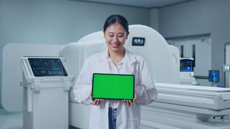 asian female doctor smiling and showing green screen tablet while standing with mri machine in the hospital