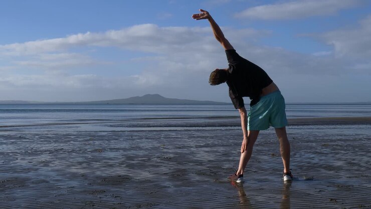 Giovane uomo in forma che fa stretching sulla spiaggia ad Auckland, Nuova Zelanda, al mattino con l'isola di Rangitoto in lontananza