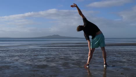 Hombre-Joven-En-Forma-Que-Se-Extiende-En-La-Playa-En-Auckland,-Nueva-Zelanda-En-La-Mañana-Con-La-Isla-Rangitoto-En-La-Distancia