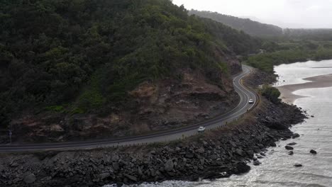 Captain-Cook-Highway-aerial-with-white-car-and-rocky-coast,-Far-North-Queensland,-Australia