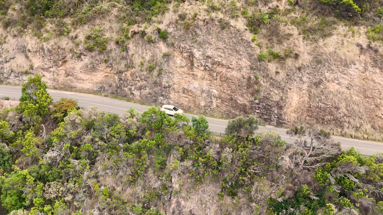 Drone captures a car navigating the scenic Great Ocean Road in Lorne, Australia, surrounded by lush greenery and coastal cliffs