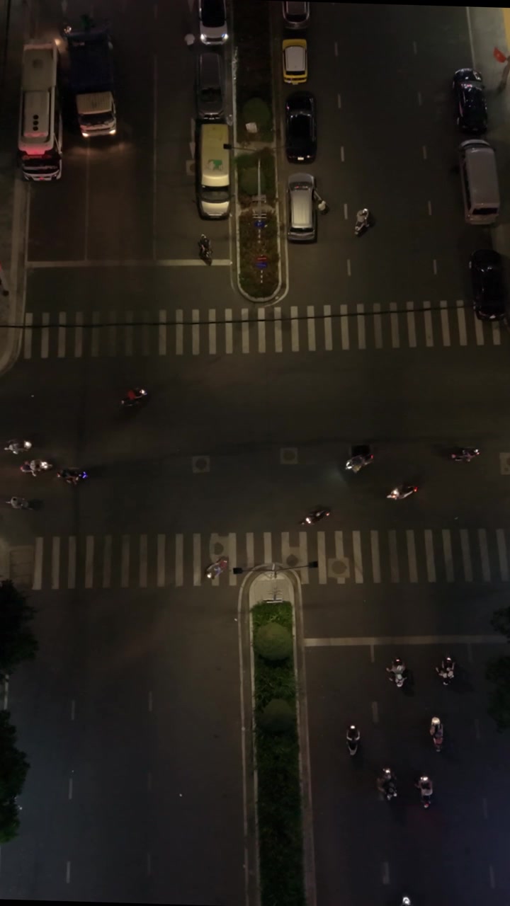 Vertical screen Close Aerial Of Scooters Crossing Central Can Tho Road At Night, Clustered Riders And Delivery Couriers Wait And Weave Through Crosswalks Under Streetlights, Asian Helmeted