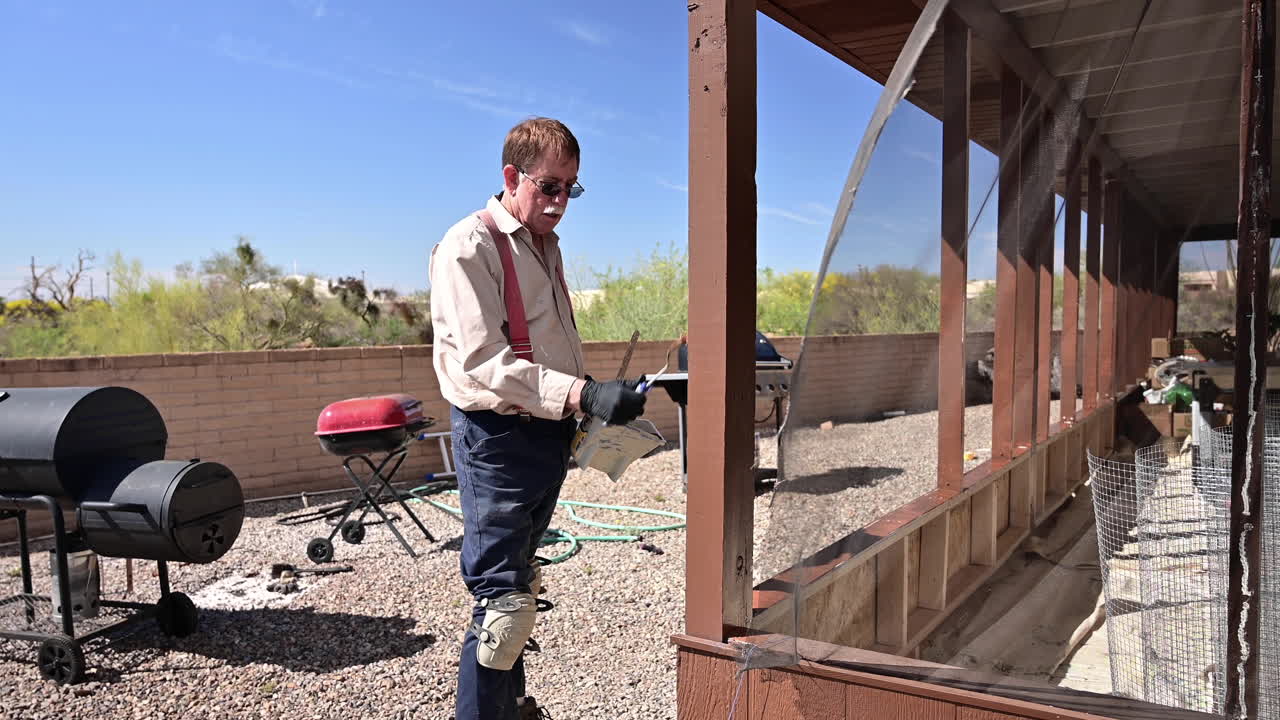 anciano pintando el exterior de la casa usando un cepillo de pintura en un día soleado con el patio trasero del desierto en segundo plano