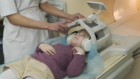 doctor radiologist puts coil on the head of the patient little girl to make an mri scanning of brain, head, neck, girl lies on automatic table, using modern equipment