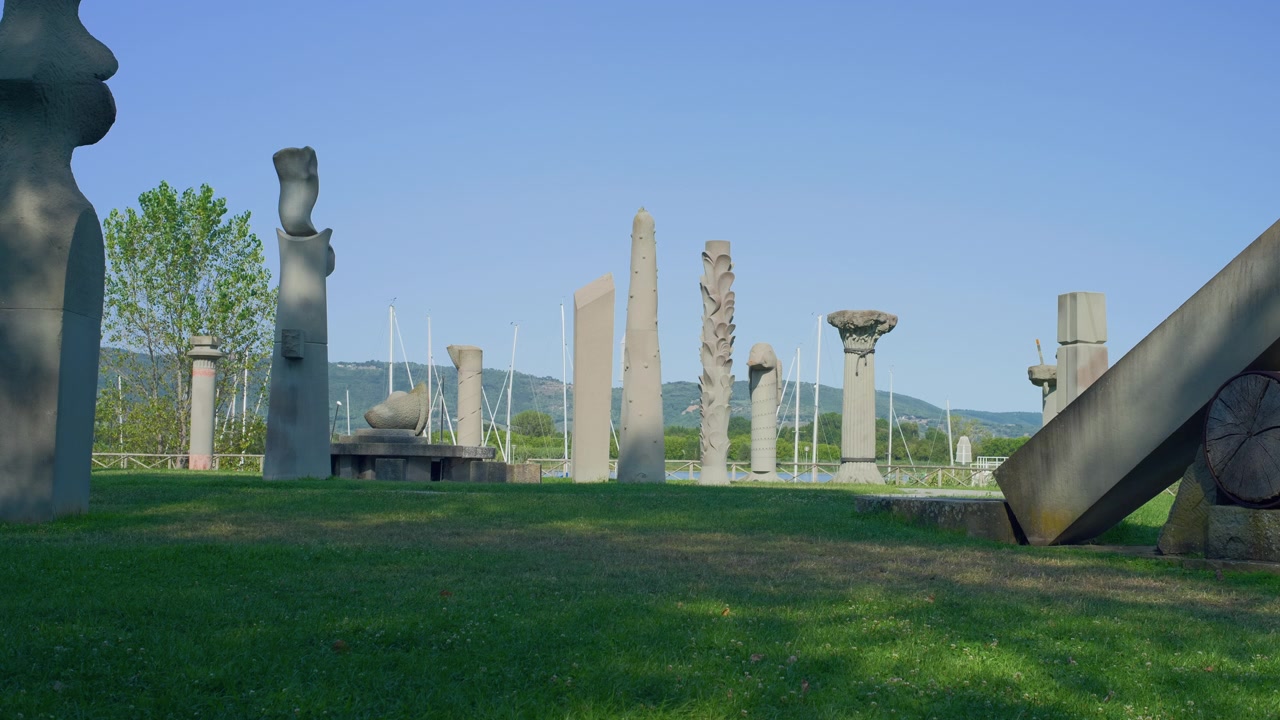 Monumen pilar batu taman patung berdiri di atas rumput hijau dekat danau di bawah langit biru yang cerah. Pameran seni terbuka kontemporer di hari yang cerah, tampilan tripod statis