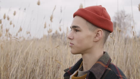 close up view of a teenage boy with orange beanie and plaid coat looking at side in a wheat field, then looks at camera and smiles