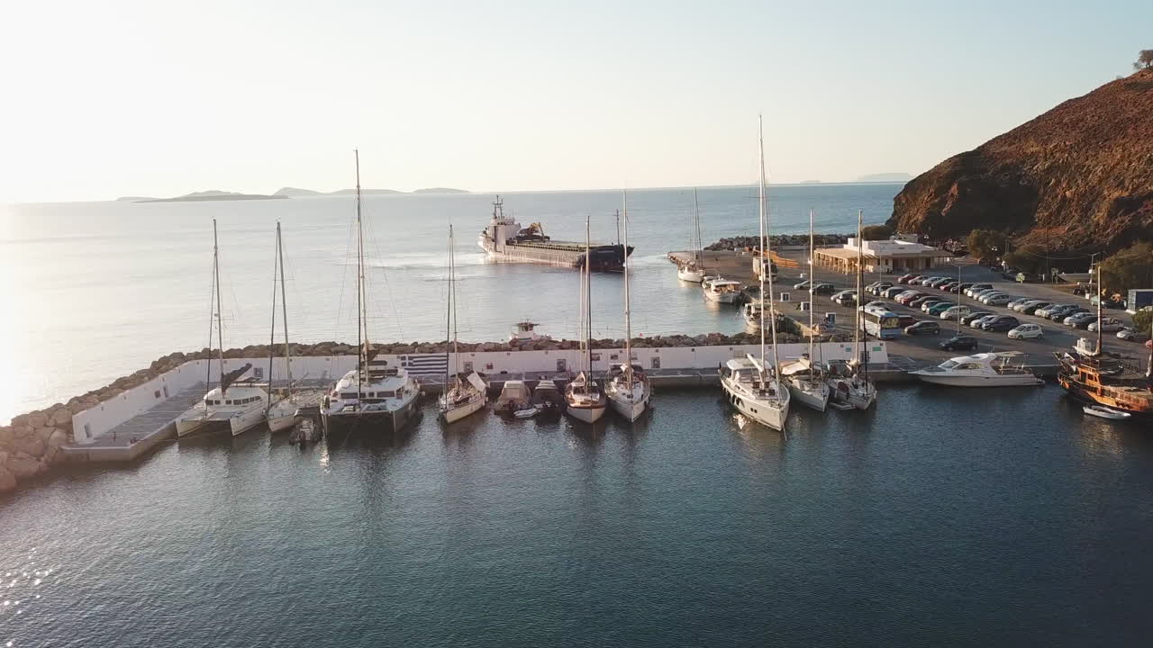 Greek Island, Astypalea's harbor view on a sunny day, drone footage starts from left slides to right and zooms in to the sail boats and behind the town quay a big vessel is trying to dock to the port
