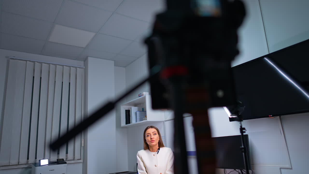 Beautiful confident Caucasian brunette lady in white jacket speaks emotionally on camera. Low angle view at the professional camera recording blogger's speech.