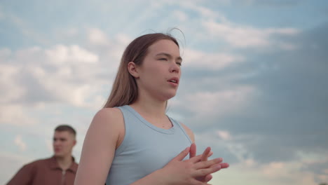 young woman standing in open field adjusts hair and takes deep breath, eyes closed in calm moment, boyfriend blurred in background under pastel cloud sky