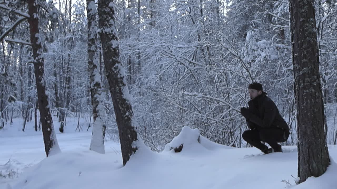 Man with black clothing squats and rubs his hands together indicating that he is cold in this winter white snow covered forest. He stands up and jumps around trying to get warm during this cloudy day.