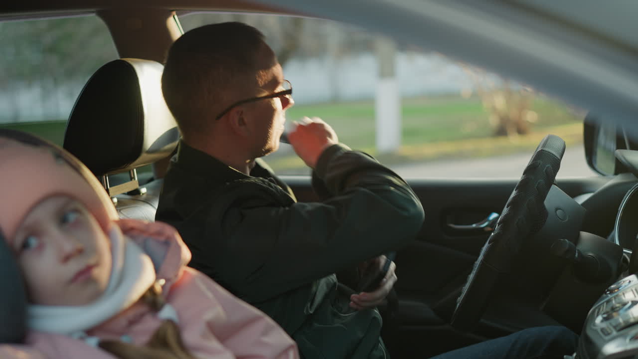 초록색 재을 입은 남자가 운전자 좌석에 앉아 전화를 들고 안전벨트를 벗고 있다. 분홍색 뜨개질 모자와  스카프를 입은 어린 소녀가 창밖을 바라보며 평화롭게 앉아 있다.