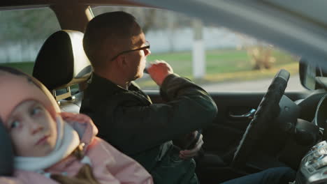 a man wearing a green jacket sits in the driver's seat, holding a phone and having just unbuckled his seat belt. a young girl in a pink knit hat and white scarf sits peacefully, looking out the window