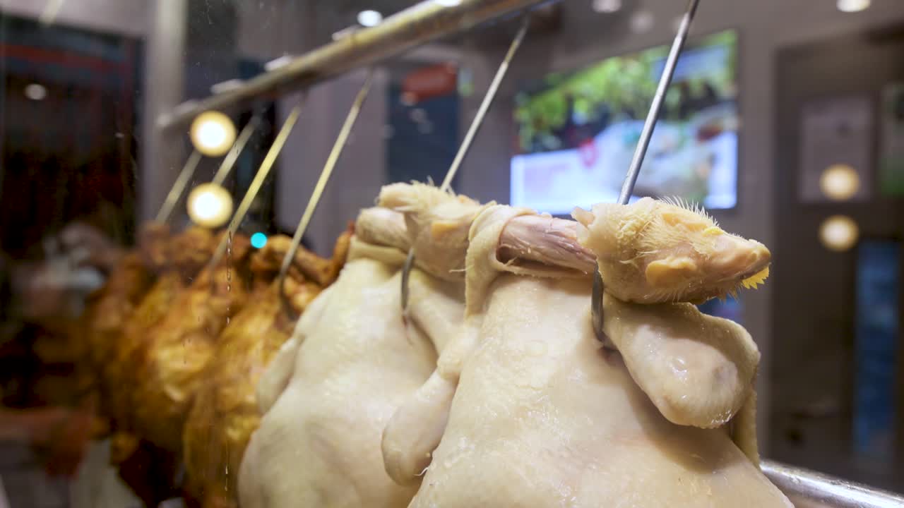 Whole steamed chickens hang on metal hooks, draining juices into a bowl below in a brightly lit commercial kitchen with shallow depth of field