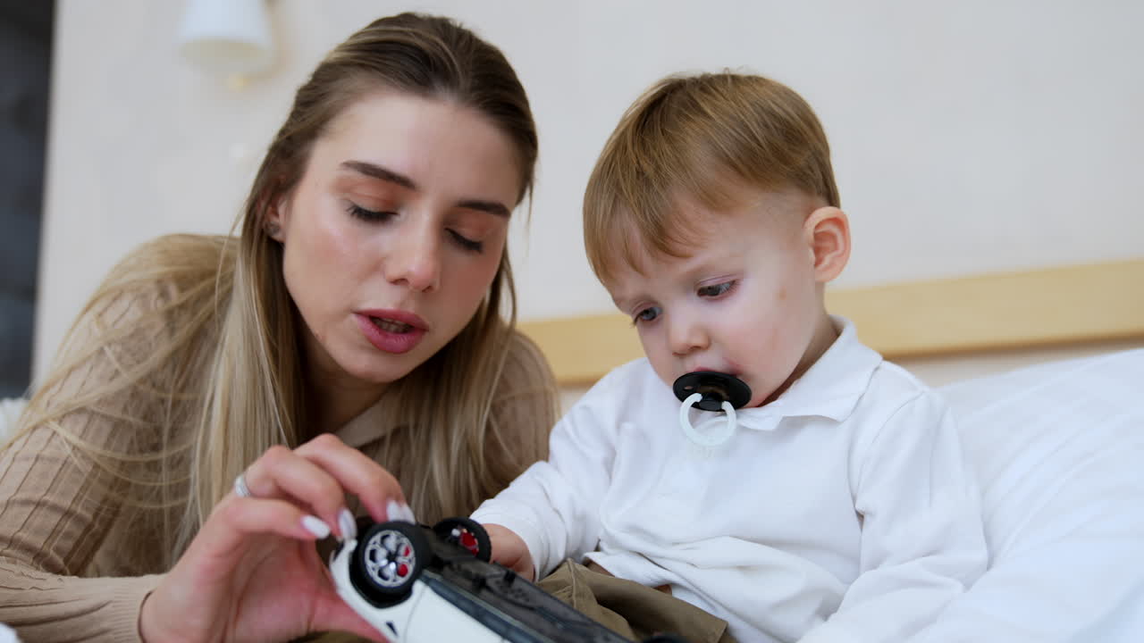 Blonde mother and cute baby boy with pacifier sit on the bed. Mom and son are holding a toy car. Close up.