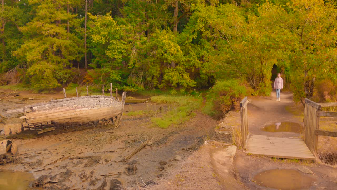 A woman strolls along a scenic hiking trail surrounded by vibrant trees and an old boat by the water's edge