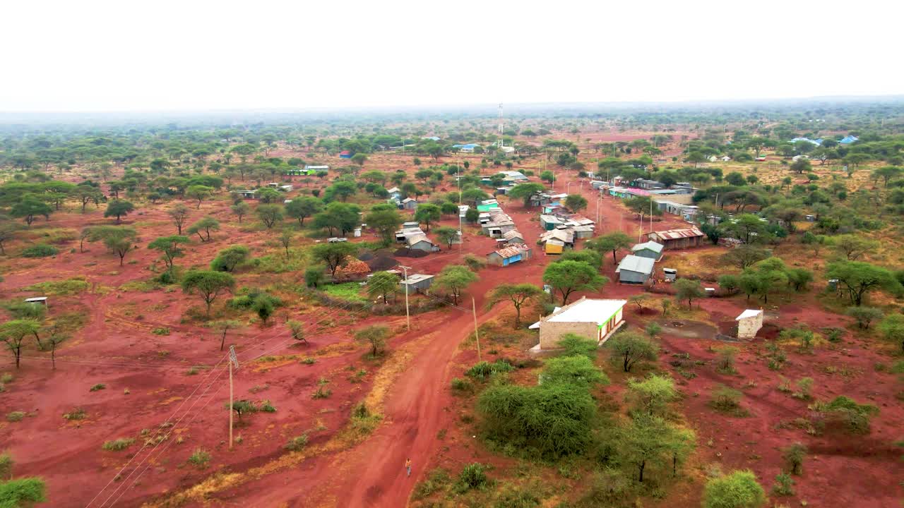 Drone aerial view of red dirt road leading to rural shopping centre surrounded by semi arid landscape with acacia trees in Kajiado Kenya