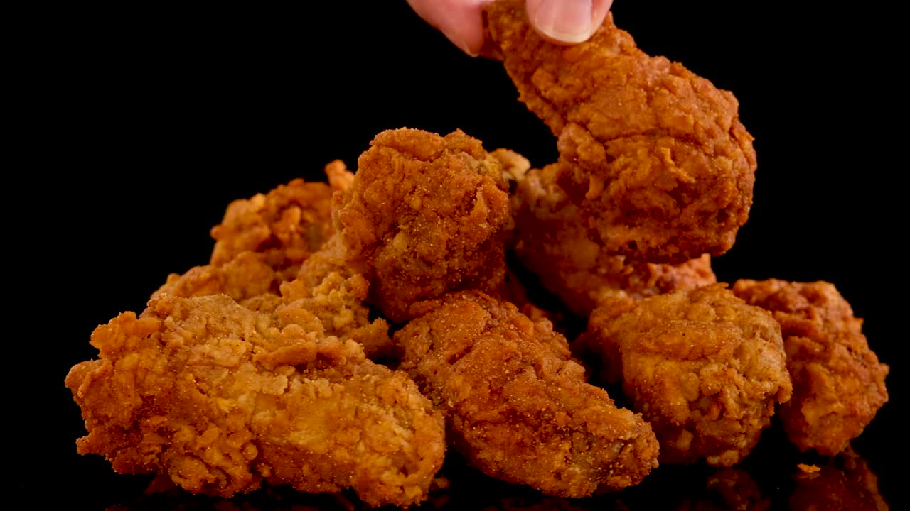 A hand slowly picks up and replaces spicy fried chicken pieces from a pile, shot in slow motion with dramatic lighting and a black background