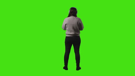 vista trasera de una mujer joven con auriculares inalámbricos transmitiendo música desde un teléfono móvil contra la pantalla verde del estudio 1