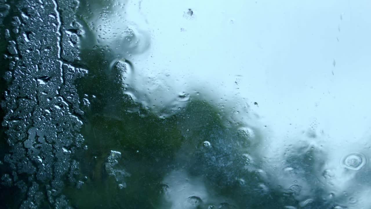 Heavy raindrops fall on window with blurry view of garden in background