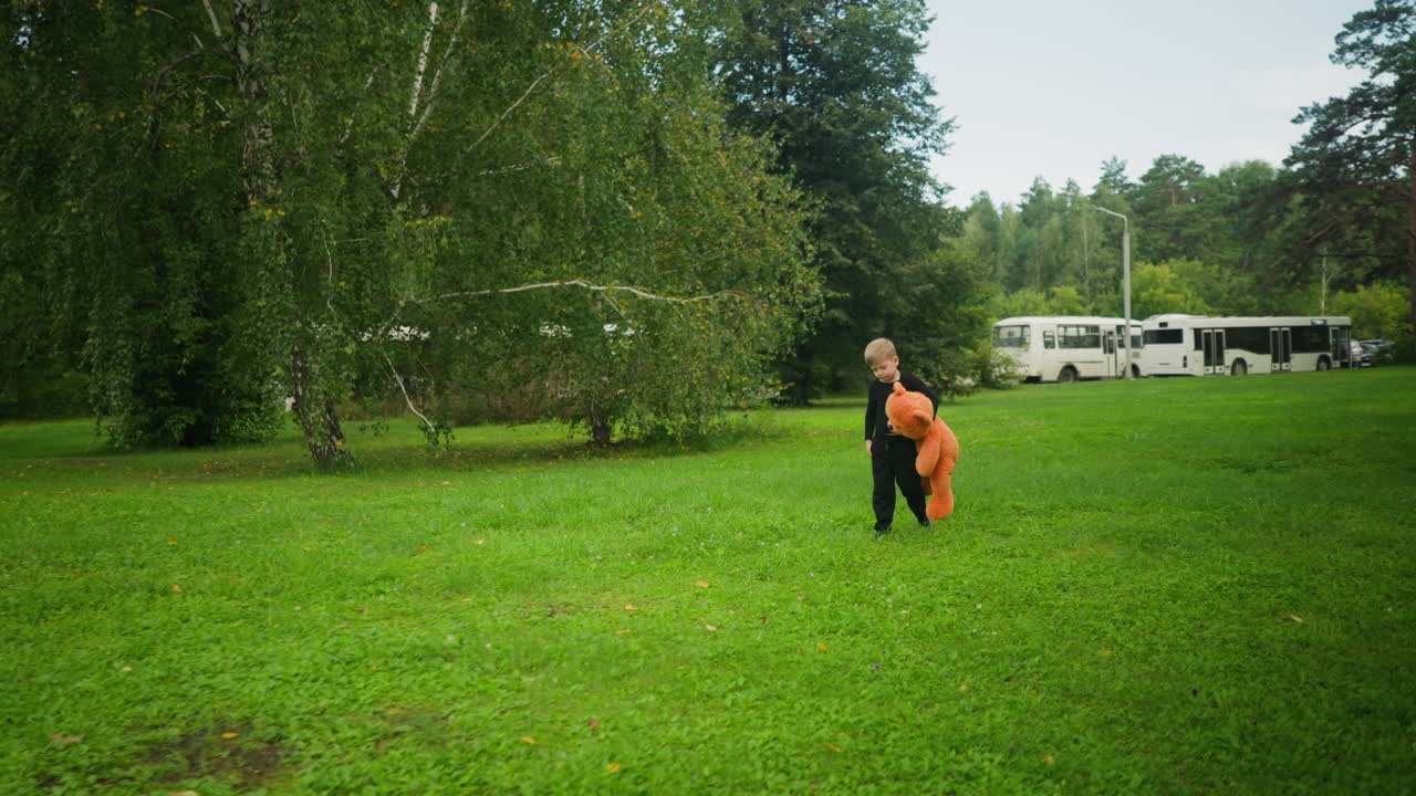 Little boy walking alone through green field holding large teddy bear with parked buses and moving car visible in distant background under calm sky near tree lined area