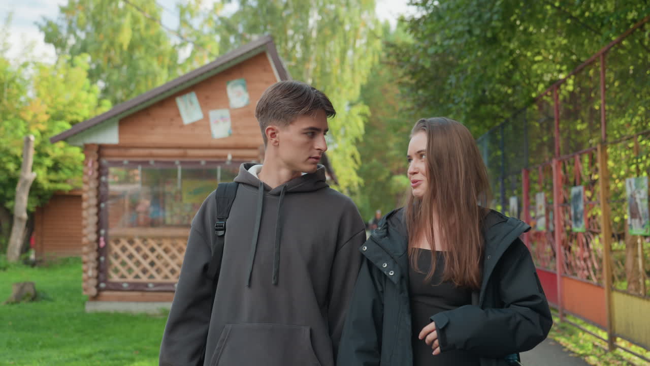 Coppia caucasica adolescente che cammina in un parco accanto a una baita in legno, con zaini e felpe con cappuccio, conversazione animata con sorrisi giocosi, il ragazzo ascolta mentre la ragazza gesticola, la scena suggerisce studenti che si dirigono verso l'estate