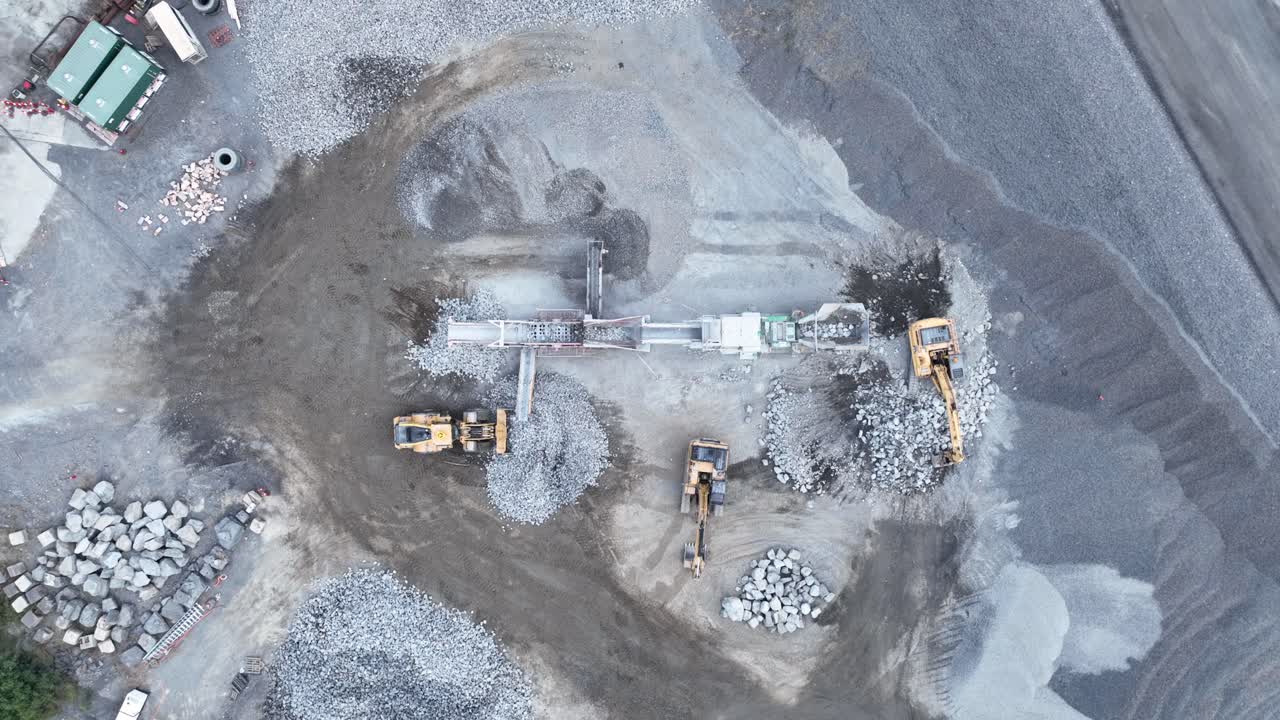 Drone stationary shot of bob cat and mining excavator moving rocks, with conveyor belt sorting rocks with rubble into piles in quarry mine