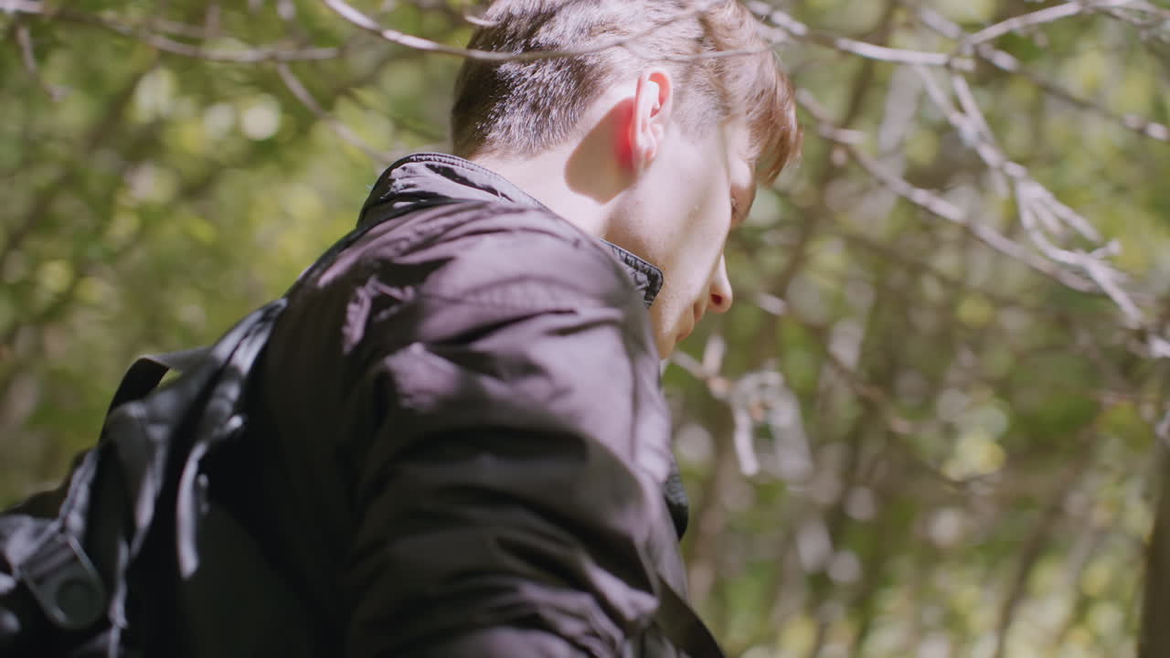 Soldier in dark jacket stands in forest, turns to walk a few steps, then pauses while holding thin tree and bends forward to observe carefully ahead, surrounded by sunlight and dense foliage