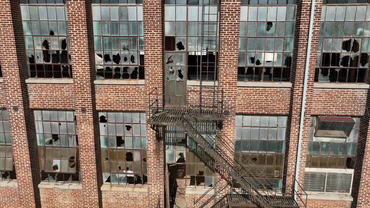 Vista ravvicinata della facciata di un vecchio edificio industriale in mattoni con finestre in frantumi e una scala antincendio metallica rustica, che trasmette degrado urbano e architettura storica