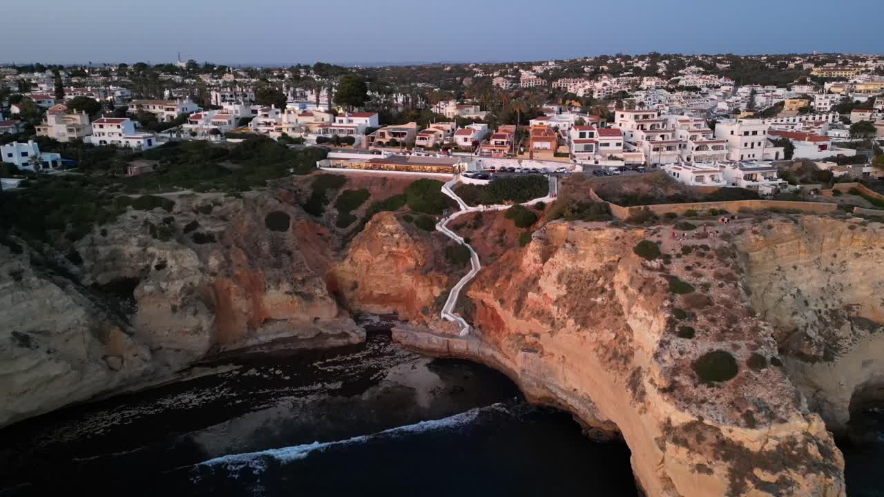 드론이 바다 위에서 카르보에로 알가르베의 아름다운 절벽으로 날아가며, 포르투갈에서 해가 지는 동안 도시가 많은 집들과 함께 보입니다.