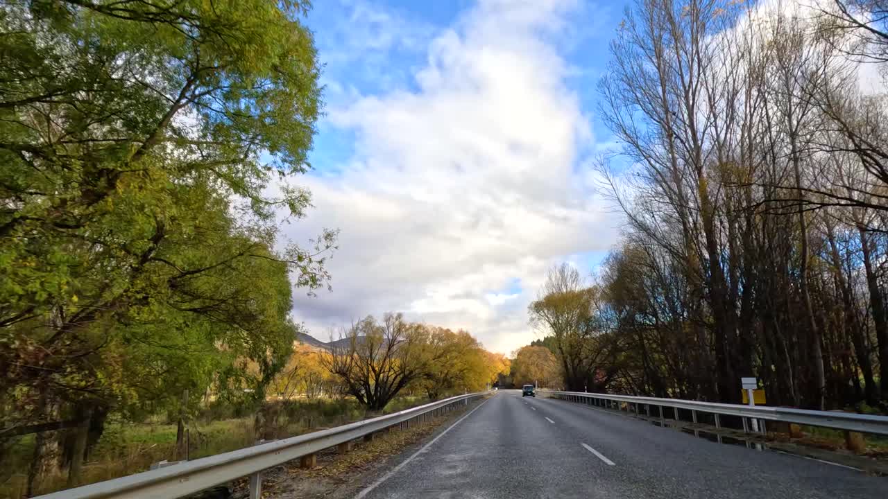 A serene drive through Wanaka, New Zealand, showcasing vibrant autumn foliage under a partly cloudy sky
