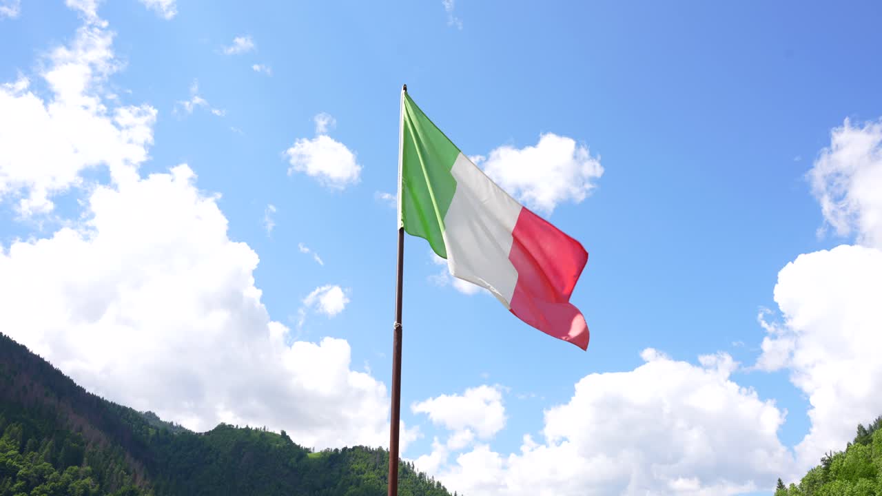 Bandiera italiana contro cielo blu con nuvole bianche, montagne sullo sfondo