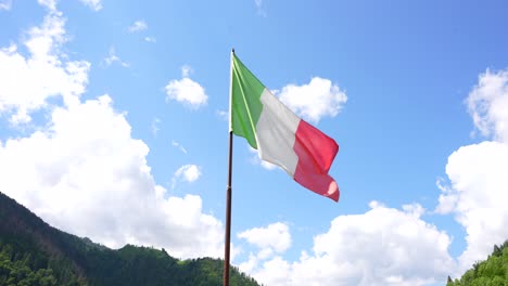 bandera italiana contra el cielo azul con nubes blancas, montañas en el fondo