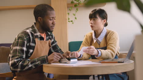 two multiethnic coffee shop owners sitting at table and doing accounting 2