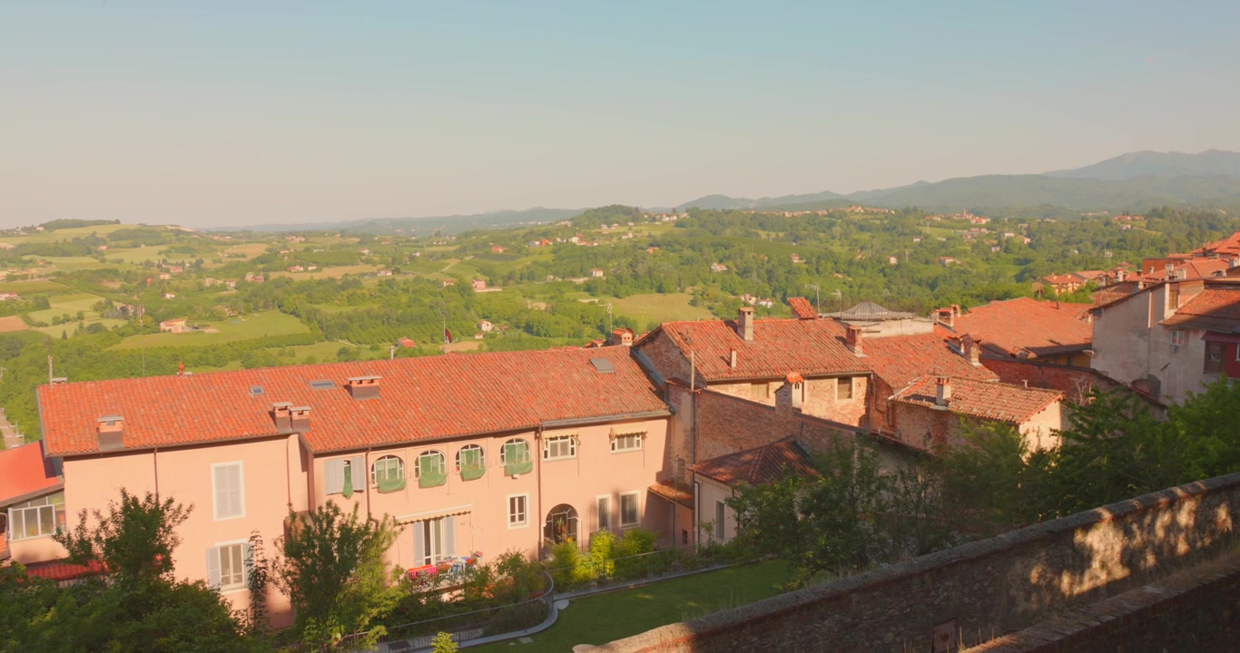 Vista dall'alto della luce calda del pomeriggio che illumina i tetti in terracotta e i giardini della città di Mondovì, con le dolci colline agricole del Piemonte e le montagne che si estendono in lontananza, Italia