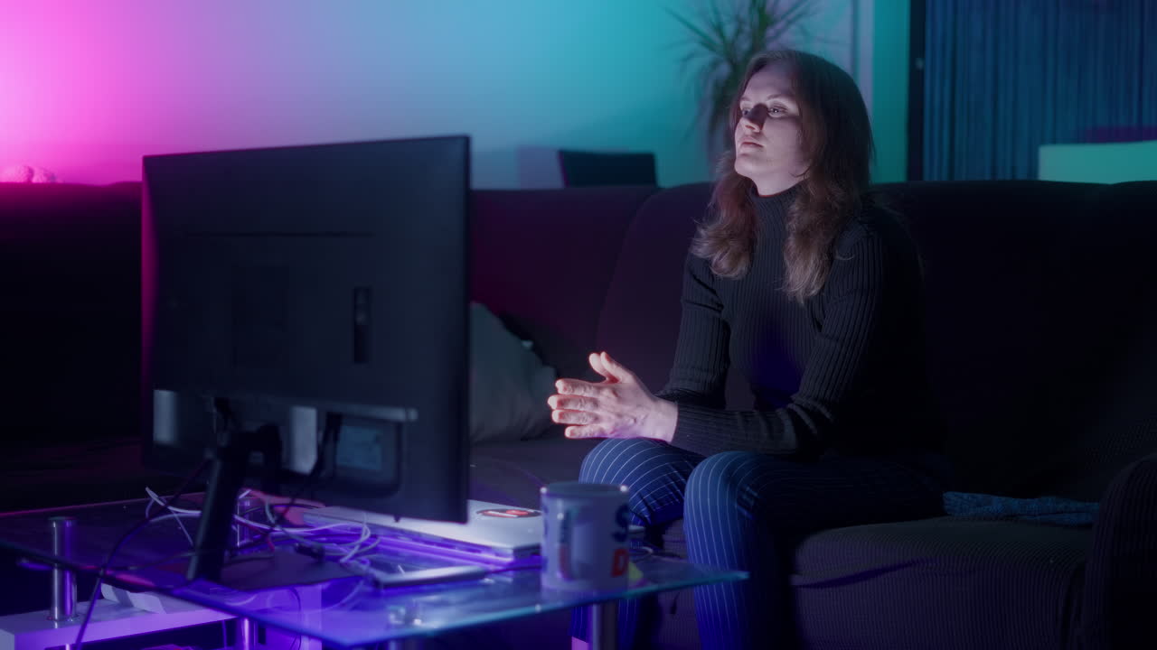 disparo medio de una mujer blanca joven viendo la televisión en su sofá negro por la noche en casa en una sala de estar iluminada cyber punk en cámara lenta