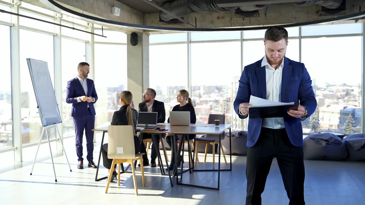 Serious entrepreneur in suit in office room. Business people planning new blueprint together. Businessman stands alone holding folder with papers.