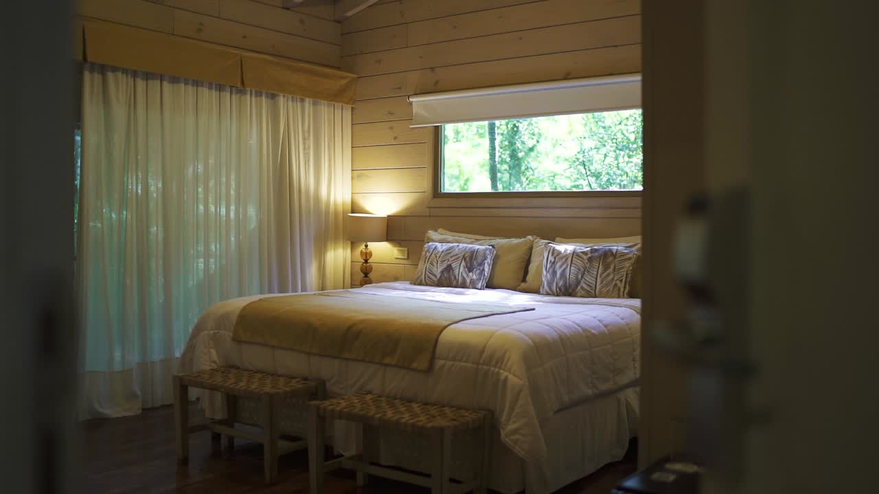 A slow panning shot moves through the doorframe of a luxury hotel room in the jungle, revealing a large kingsize bed surrounded by elegant decor and natural beauty