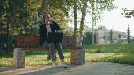 freelancer takes break drinking water while working outdoors with laptop on lap, seated on wooden bench in park with sunlight filtering through trees, surrounded by lush greenery