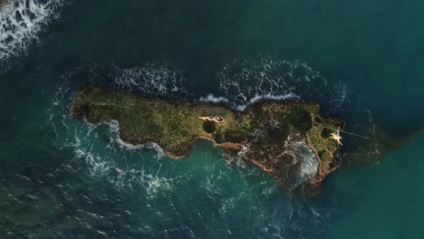 picturesque summertime aerial scene of caucasian woman laying down outstretched on rocky outcropping in beautiful blue ocean sea water with waves crashing by sandy beach, directly above rising