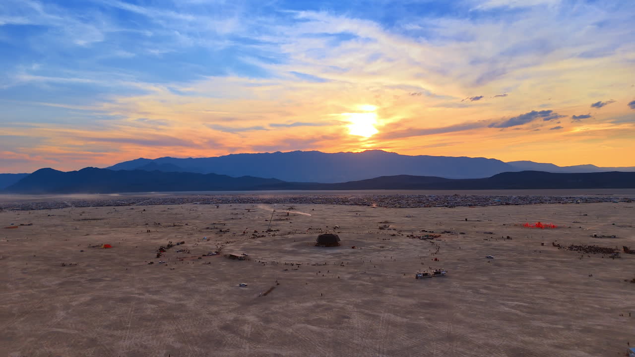 Aerial panoramic view of the Burning Man city and installations glowing under a colorful sunset sky
