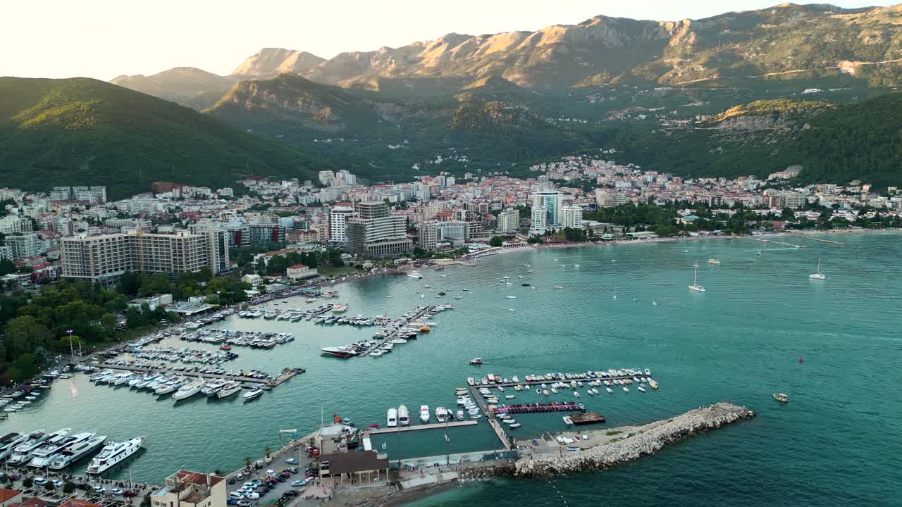 Panoramic aerial view of Budva city. Famous travel destination in Montenegro. Sunset point, warm colours on mountains peacks. Port of Budva. Drone going forward descending.