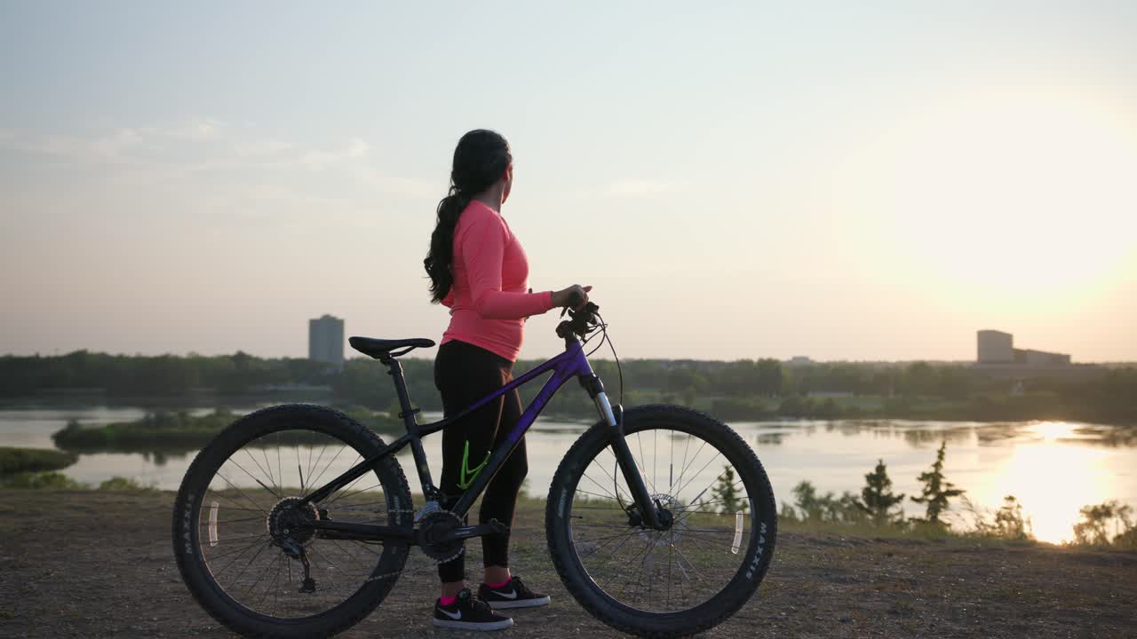 Woman stands with her bike while she takes off her helmet and looks out over a lake