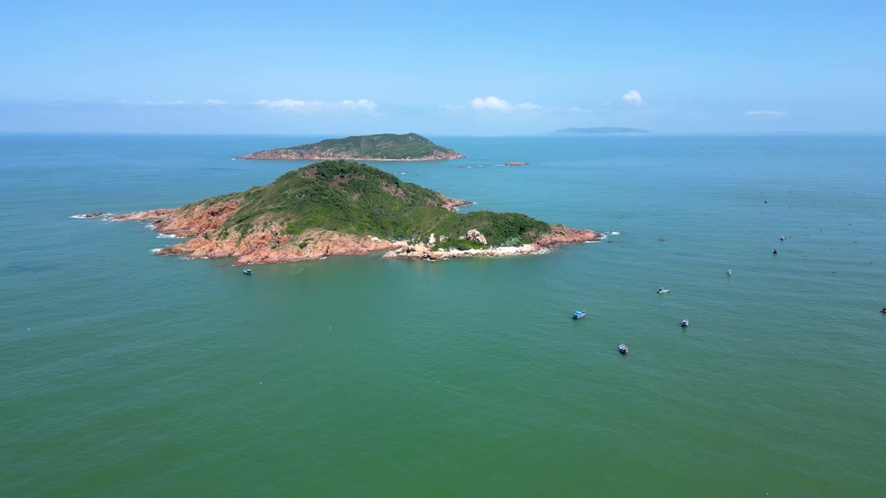 Drone flight toward rock island with vegetation and fishing boats in ocean
