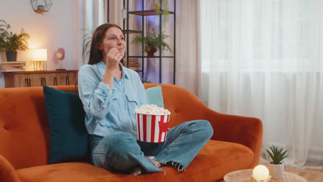 joven emocionada sentada en el sofá comiendo palomitas de maíz y viendo un interesante juego de serie de televisión