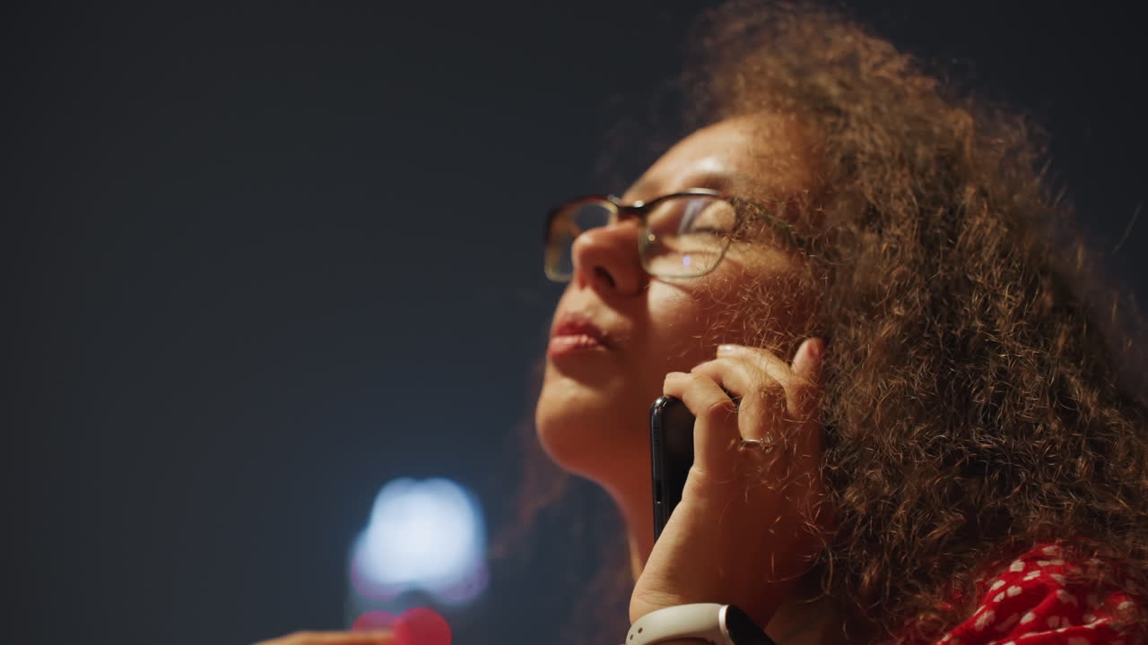 Femme en ville à minuit au téléphone, pensive, cheveux bouclés et lunettes encadrées par un bokeh bleu froid, main près du visage et lueur néon lointaine, humeur introspective, conversation discrète cherchant du réconfort, contemplative.