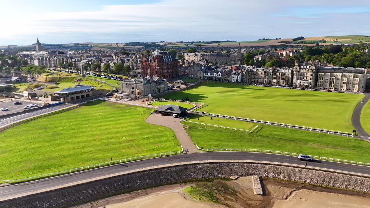 Drone footage glides above lush green golf course, Victorian brick architecture, and coastal landscape in bright daylight at St Andrews, Scotland