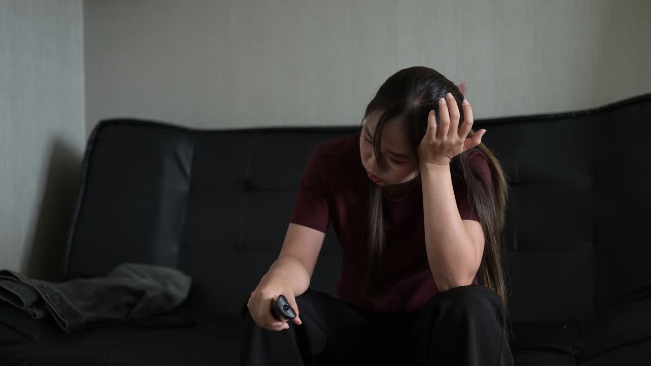 Stressed Asian woman sitting on sofa and watching television holding remote control while getting bad news