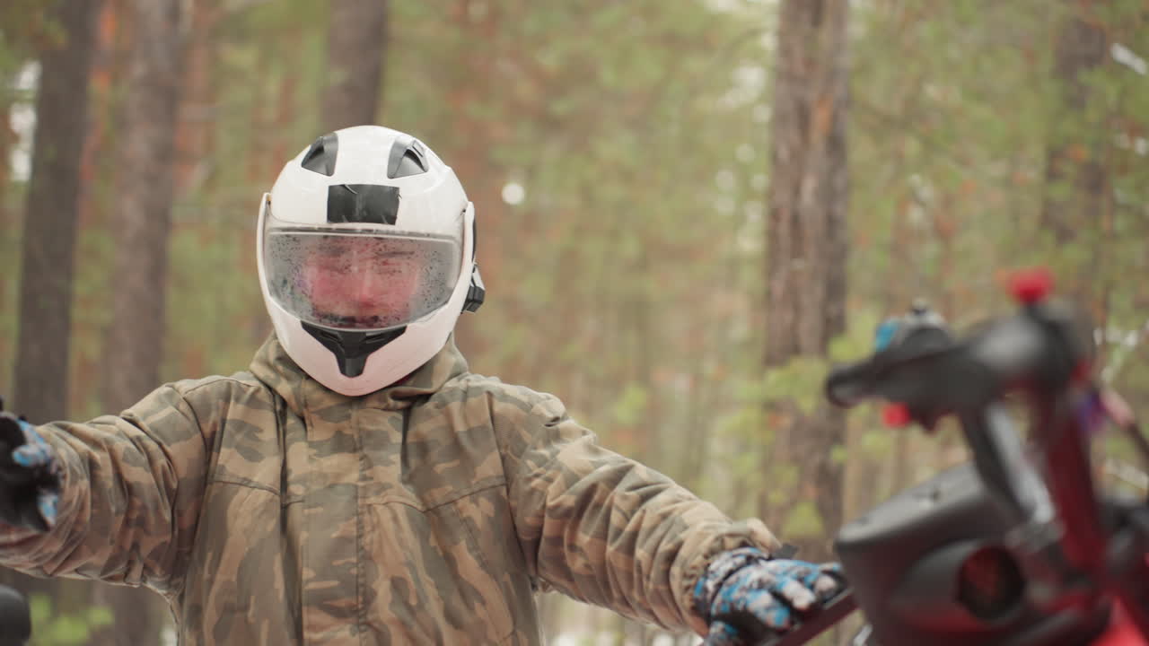 Un pilota a figura intera indossa il casco dell'ATV in mezzo a una pineta. Tuta mimetica e guanti, visiera e cinturino del casco bianco ben assicurati. Il pilota sale sul manubrio e testa i comandi, cogliendo una tranquilla anticipazione.