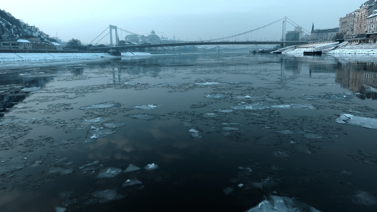 Frozen Danube River in Budapest with drifting ice and Liberty Bridge visible in the background. Cold winter atmosphere, overast sky, historic city waterfront and calm urban landscape
