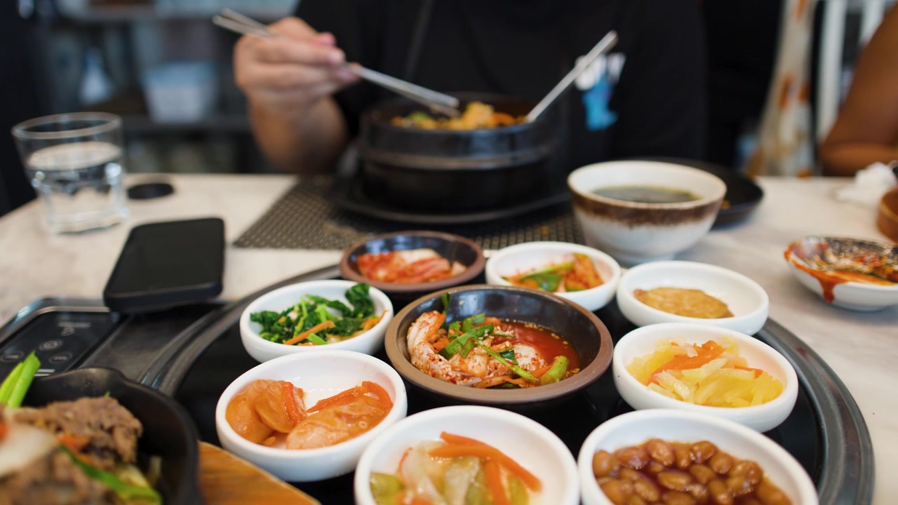 Seseorang menggunakan sumpit logam untuk makan berbagai lauk Korea dan sup sayuran pedas
